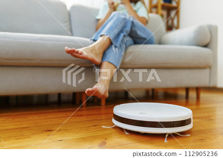 Person relaxing on sofa with feet up while robotic vacuum cleaner works on floor Person relaxing on sofa with feet up while robotic vacuum cleaner works on floor 112905236