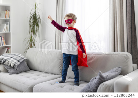 Child in superhero costume with red cape and mask standing on a sofa Child in superhero costume with red cape and mask standing on a sofa 112905244