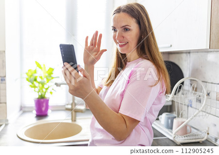 Woman waving at phone during a video call in a bright kitchen Woman waving at phone during a video call in a bright kitchen 112905245