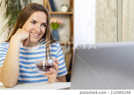 Smiling woman using laptop and holding cup of tea. Smiling woman using laptop and holding cup of tea. 112905246