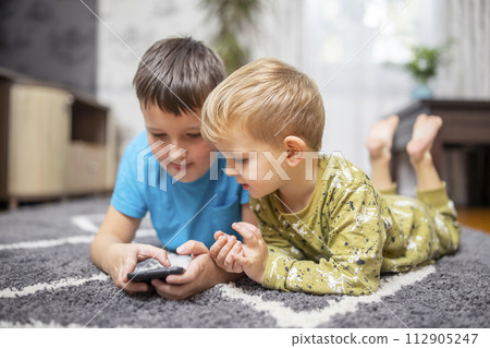 Two young boys lying on carpet using smartphone together Two young boys lying on carpet using smartphone together 112905247