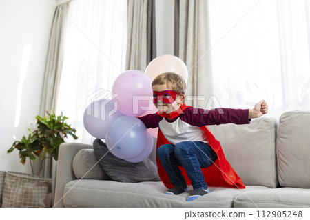 Boy in superhero costume with red cape and mask playing with balloons. Boy in superhero costume with red cape and mask playing with balloons. 112905248