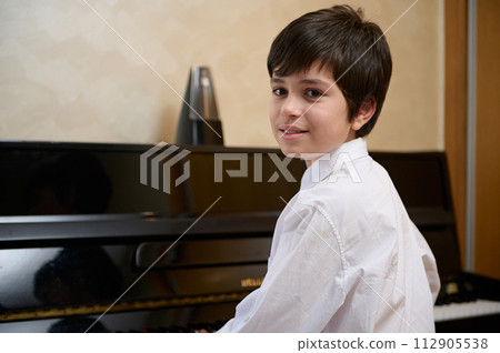 Close-up portrait of a multi ethnic teen boy, little pianist musician smiling looking at camera, sitting at piano forte Close-up portrait of a multi ethnic teen boy, little pianist musician smiling looking at camera, sitting at piano forte 112905538