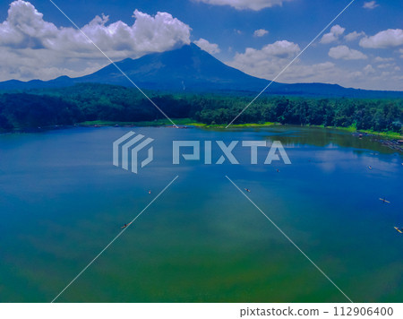 An aerial view of a clear blue lake with a dramatic, clouds capped mountain in the background. Evergreen trees line the shore. An aerial view of a clear blue lake with a dramatic, clouds capped mountain in the background. Evergreen trees line the shore. 112906400