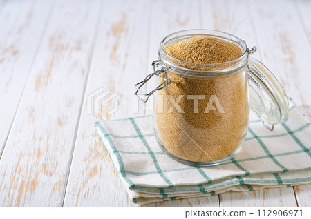 Uncooked dry amaranth grains in glass storage jar on a white wooden table. 112906971