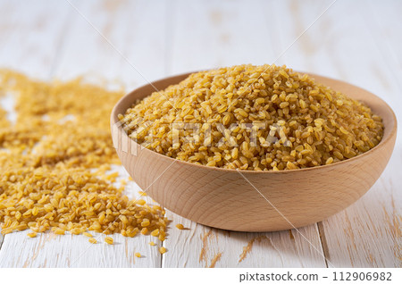 Raw bulgur are scattered out of the wooden bowl on a light table, selective focus. Raw bulgur are scattered out of the wooden bowl on a light table, selective focus. 112906982