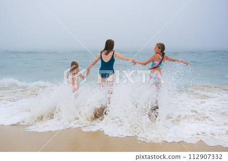 Mother and children playing on the ocean beach. Family enjoying the ocean. Mother holds girls's hands and they all look at the ocean together. Mother and children playing on the ocean beach. Family enjoying the ocean. Mother holds girls's hands and they all look at the ocean together. 112907332
