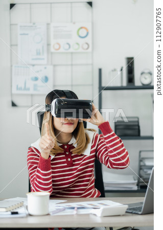 Professional in a red and white striped sweater interacts with a virtual environment using a VR headset at her office desk. 112907765