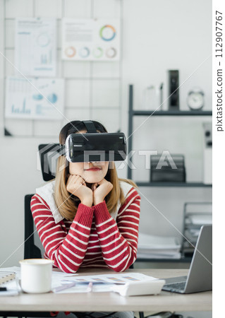 Contemplative young woman with a VR headset sits at her office workspace, exploring virtual environments. 112907767