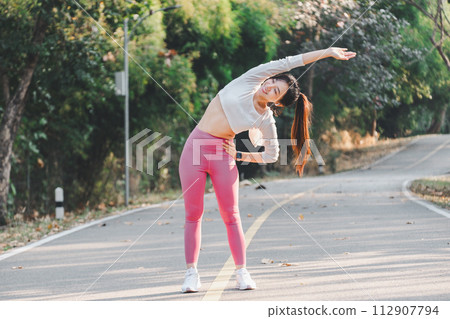 Athletic woman performs a side stretch on a serene park road, engaging in a healthy fitness routine. Athletic woman performs a side stretch on a serene park road, engaging in a healthy fitness routine. 112907794