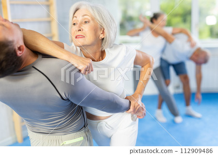 Senior woman in pair with middle-aged coach train Krav Maga and self-defense skills. 112909486