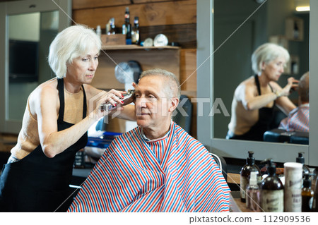 Female barber making haircut with clippers to elderly client in barbershop Female barber making haircut with clippers to elderly client in barbershop 112909536