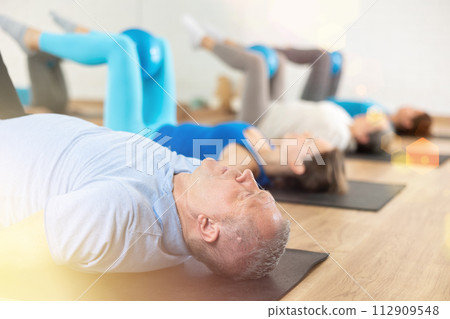 People of different ages pressing core and clutching mini Pilates balls between legs during Pilates training together 112909548