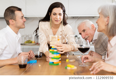 Group of mature spouses and son with girlfriend is playing board game with wooden blocks in kitchen Group of mature spouses and son with girlfriend is playing board game with wooden blocks in kitchen 112909550