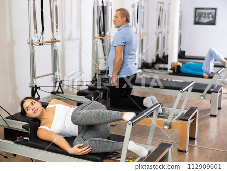 Focused young woman in activewear performing Pilates exercise for legs and back, training on bed equipment in gym Focused young woman in activewear performing Pilates exercise for legs and back, training on bed equipment in gym 112909601