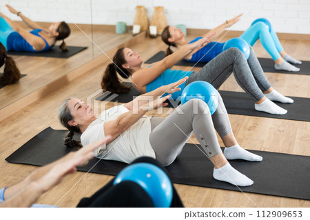 People of different ages pressing core and clutching mini Pilates balls between legs during Pilates training together 112909653