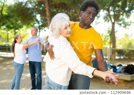 Exited older and middle-aged diverse people enjoying latin dances breaking into pairs on sandy area between trees 112909696
