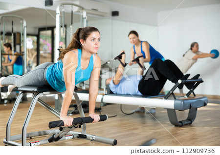 Young woman practicing pilates on pedal fitness chair in sports hall Young woman practicing pilates on pedal fitness chair in sports hall 112909736