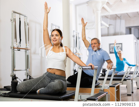 Active young woman stretching upper body practicing Pilates exercise in studio with elderly people 112909815