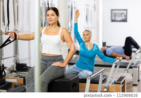 Calm smiling young woman in sportswear pulling hand in band from Pilates reformer machine during workout in small studio Calm smiling young woman in sportswear pulling hand in band from Pilates reformer machine during workout in small studio 112909816