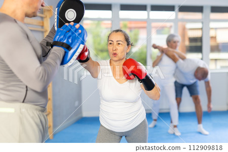Senior lady and male couch are boxing in gym, man helps student to work out force of blow. 112909818