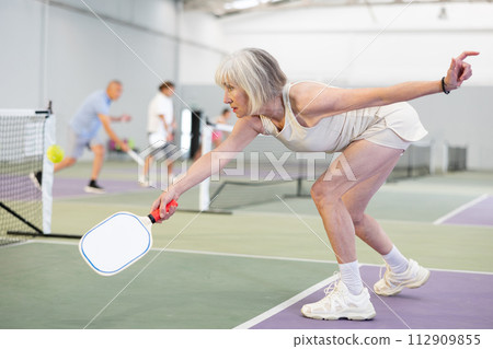 Positive elderly woman playing pickleball on indoor court 112909855