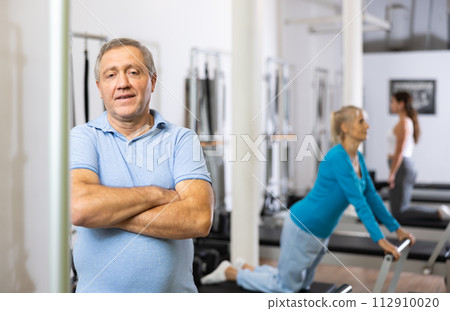 Portrait of smiling elderly male pensioner in activewear posing in modern Pilates fitness studio Portrait of smiling elderly male pensioner in activewear posing in modern Pilates fitness studio 112910020