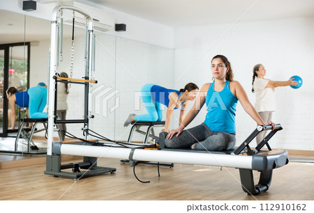 Concentrated young female in active wear training and using Pilates performer bed in gym 112910162