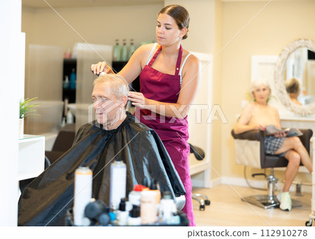 Young female hairstylist making haircut with electric trimmer to elderly client 112910278