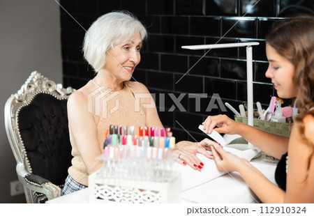 Unrecognizable girl manicure master files nails and performs grooming procedure for senior woman 112910324