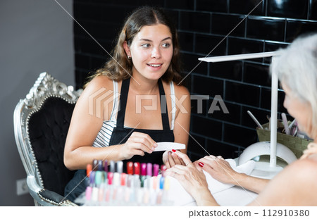 Skilled woman manicurist filing and shaping nails of female client during procedure of manicure in beauty salon Skilled woman manicurist filing and shaping nails of female client during procedure of manicure in beauty salon 112910380