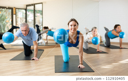 People of different ages stretching leg and arm, holding mini ball in their hands during Pilates workout in studio 112910405