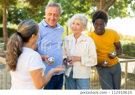 Excited multinational people listening to explanations about petanque game against trees in afternoon park Excited multinational people listening to explanations about petanque game against trees in afternoon park 112910619