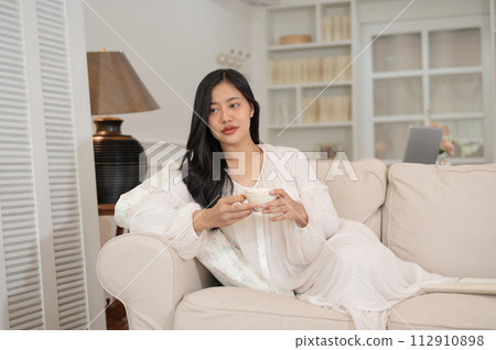 An attractive Asian woman in a long dress pajamas is sipping tea and relaxing on a sofa. 112910898