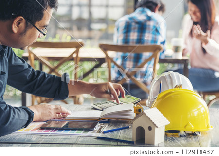 Civil construction engineer working with laptop at desk office with white yellow safety hard hat at office on construction site. Asian young man architecture project manager sitting at office on site 112918437