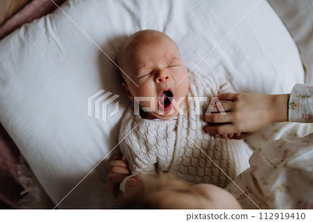 Top view of cute little baby yawning in bed, big sister holding newborn hand. Sisterly love, joy for new family member. Top view of cute little baby yawning in bed, big sister holding newborn hand. Sisterly love, joy for new family member. 112919410