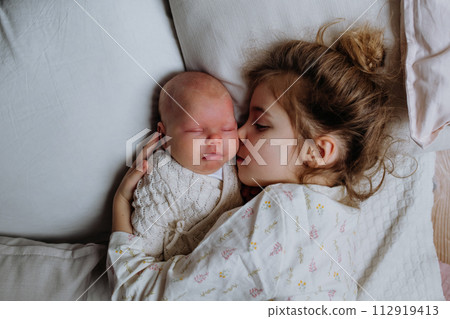 Portrait of big sister cuddling newborn, little baby. Girl lying with her new sibling in bed, closed eyes. Sisterly love, joy for new family member. Portrait of big sister cuddling newborn, little baby. Girl lying with her new sibling in bed, closed eyes. Sisterly love, joy for new family member. 112919413
