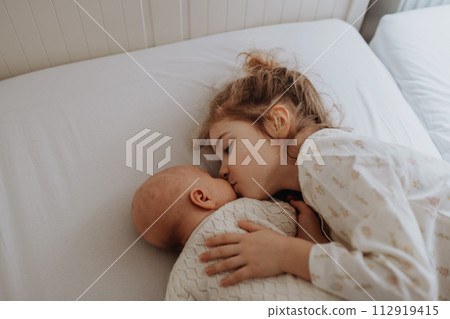 Portrait of big sister cuddling newborn, little baby. Girl lying with her new sibling in bed, kissing baby's cheek. Sisterly love, joy for new family member. Portrait of big sister cuddling newborn, little baby. Girl lying with her new sibling in bed, kissing baby's cheek. Sisterly love, joy for new family member. 112919415