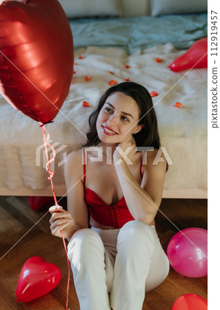 Beautiful woman sitting by bed in the middle of baloons, holding heart shaped balloon in hand. Concept of Valentine's Day, love and romance. 112919457