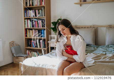 Woman at home suffering from menstrual pain, having cramps. Woman warming lower abdomen with a hot water bottle, endometriosis, and conditions causing pain in tummy. 112919476
