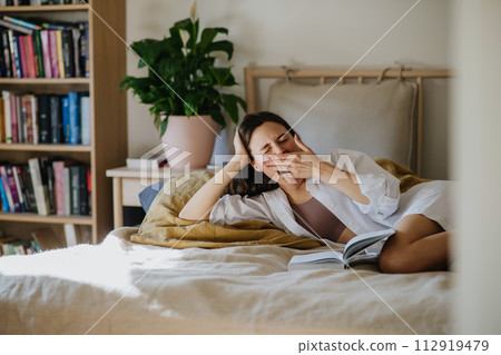Beautiful woman yawning, reading book in bed, cozy, warm and contented feeling. Freedom in story, weekend activity for single woman. 112919479