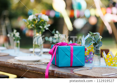 Close up of birthday gift in blue wrapping paper with pink ribbon. Summer garden birthday party in a beautiful garden. 112919527