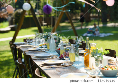 Close up shot of set table at summer garden party, grilled food. Table setting with glasses, lemonade, delicate floral and paper decoration, and bottles of summer wine. 112919539