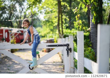 Beautiful young girl sitting on white wooden fence, helping on family farm during summer. Beautiful young girl sitting on white wooden fence, helping on family farm during summer. 112920671