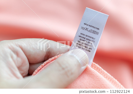 Hand hold and reading at white laundry care washing instructions clothes label on pink shirt Hand hold and reading at white laundry care washing instructions clothes label on pink shirt 112922124