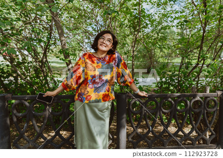 Joyful young woman in colorful blouse enjoying a sunny day in the park. Stylish asian woman enjoying a summer day while walking in a green park 112923728