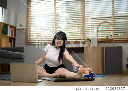 Happy young woman stretching her legs on mat and watching a tutorial on laptop Happy young woman stretching her legs on mat and watching a tutorial on laptop 112923837