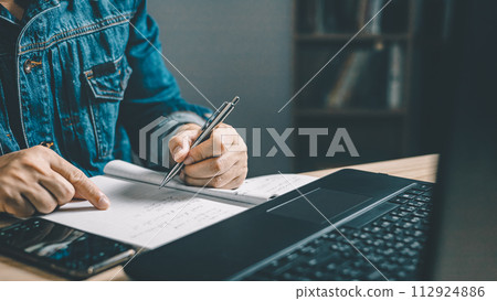 Man using smartphone and taking notes in planning. which is payment online, sits on the chair in the living room at home on a desktop. The concept of finance and online shopping. Man using smartphone and taking notes in planning. which is payment online, sits on the chair in the living room at home on a desktop. The concept of finance and online shopping. 112924886