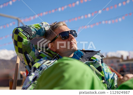 Snowboarder soaking up the sun in a lounge chair at a ski resort, surrounded by a lively atmosphere. Snowboarder soaking up the sun in a lounge chair at a ski resort, surrounded by a lively atmosphere. 112924987