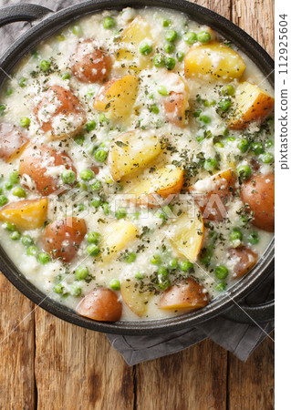 Potatoes and green peas baked in creamy cheese sauce close-up in a frying pan on a wooden table. Vertical top view 112925604